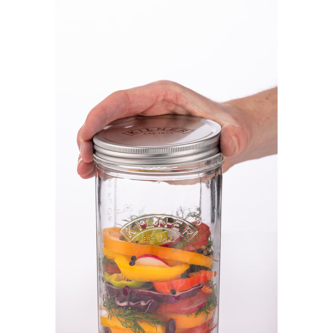 Hand holding a glass jar with a metal lid filled with sliced vegetables on a white background