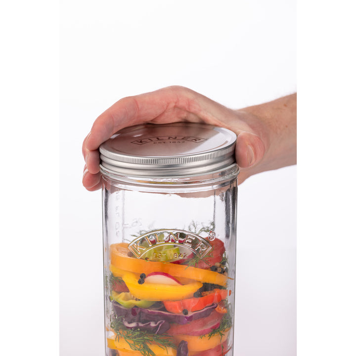 Hand holding a glass jar with a metal lid filled with sliced vegetables on a white background