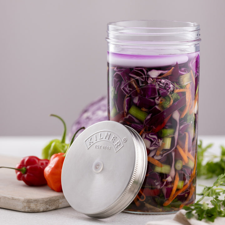 Glass jar with a purple lid filled with shredded vegetables on a white surface with vegetables around.