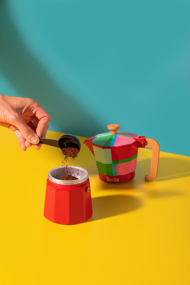 Person adding coffee grounds to a red coffee maker with a colorful teapot in the background on a yellow surface.