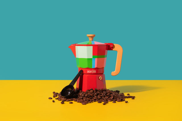 Colorful coffee maker with coffee beans and a spoon on a yellow surface with a blue background