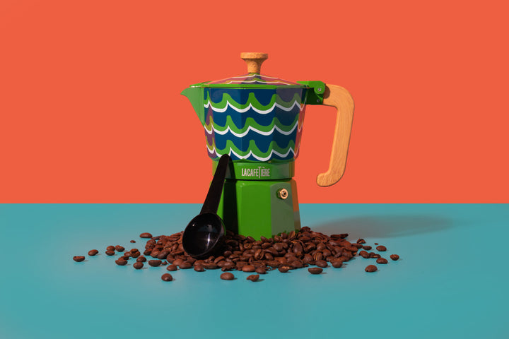 Colorful coffee percolator with wavy design on a blue surface with coffee beans and a spoon.