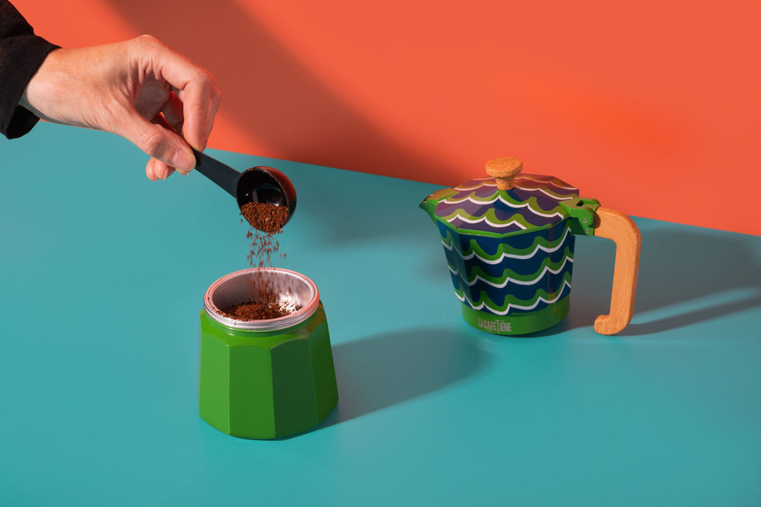 Hand pouring coffee into a green and white container with a colorful teapot on an orange and blue background.