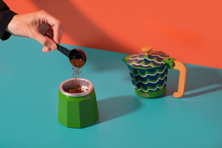 Hand pouring coffee into a green and white container with a colorful teapot on an orange and blue background.