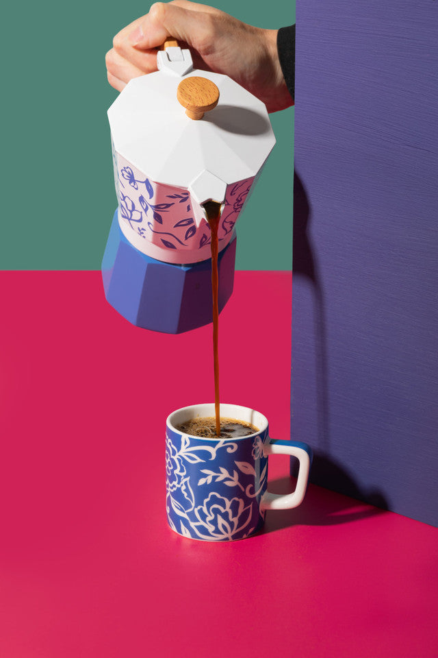 Person pouring coffee from a colorful coffee pot into a matching mug on a pink and green background.