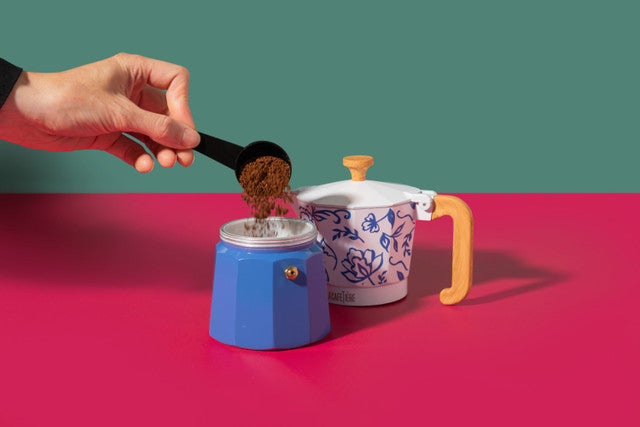Person adding coffee grounds to a blue coffee maker with a white and purple patterned pot in the background.
