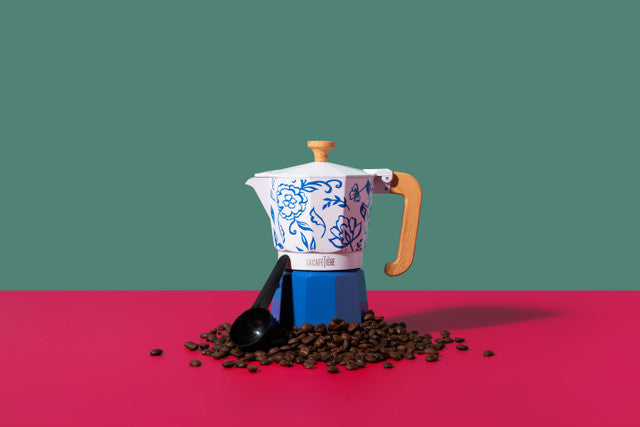 Coffee maker with floral design on a red surface with coffee beans and a black scoop.