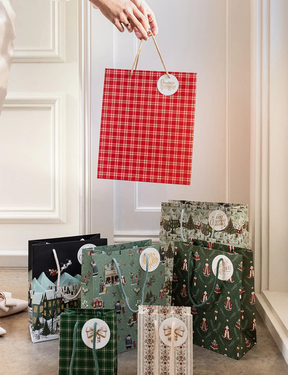 Red plaid gift bag held above other patterned gift bags on a light-colored floor.