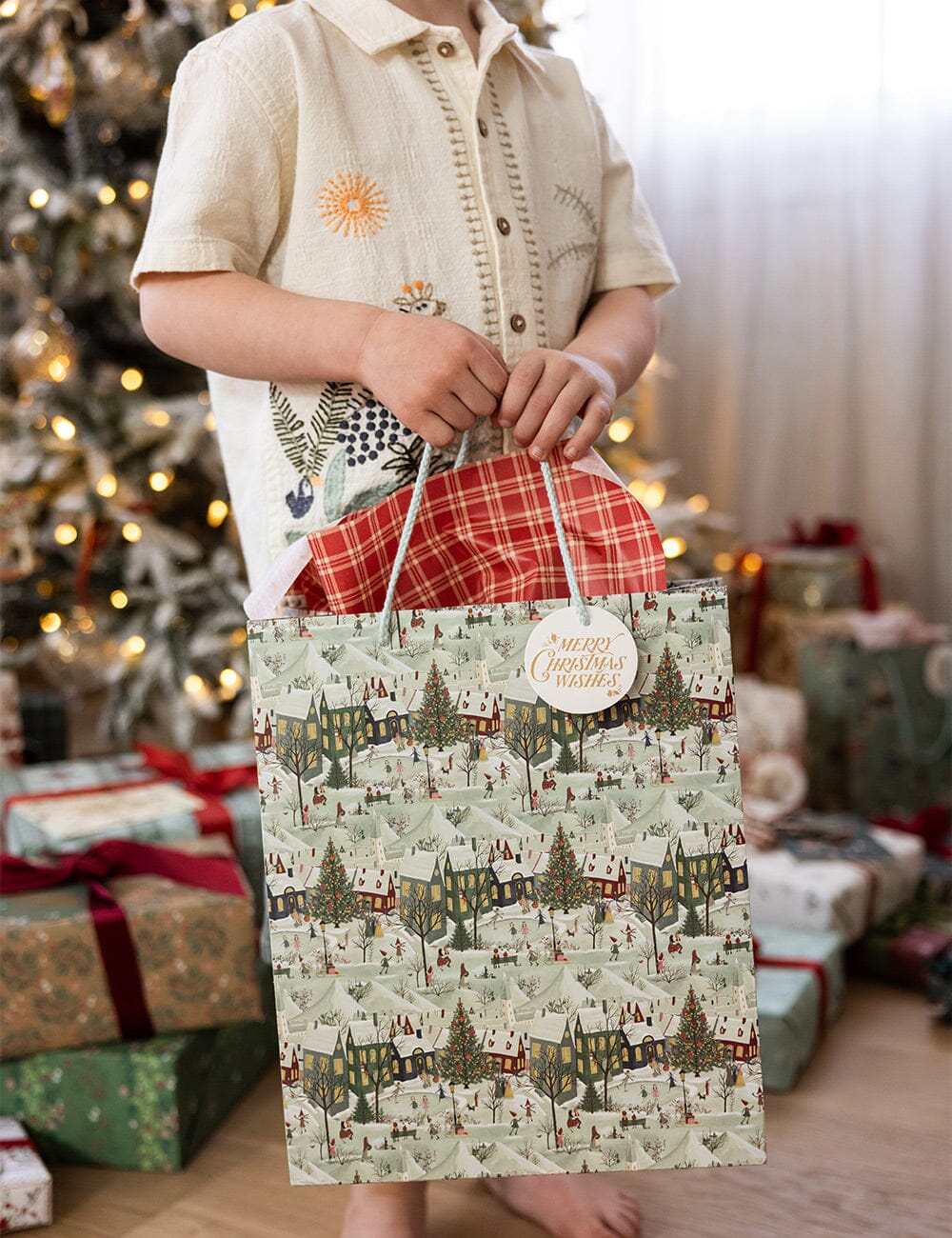 Person holding a Christmas-themed gift bag with a decorated tree and presents in the background.