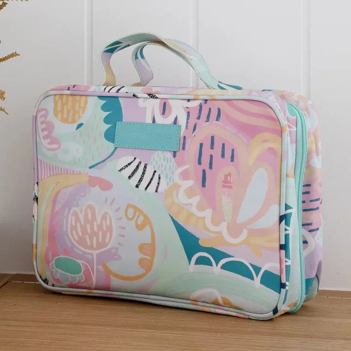 A rectangular zippered toiletry bag with a pastel multicolor abstract floral pattern, featuring a top handle and a light blue front label, resting on a wooden surface against a white paneled wall.