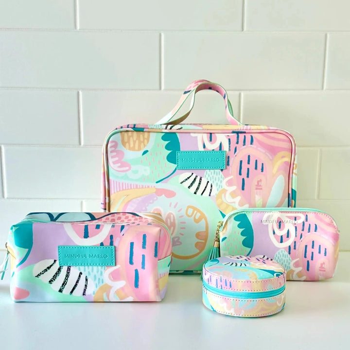 A matching set of four pastel patterned cosmetic bags and a small round zip case arranged on a white surface against a white subway tile wall. The set includes a large tote-style bag, a medium zip case, a small zip pouch, and a round round case, all featuring a colorful abstract print in pink, mint, peach, and blue with turquoise accents.