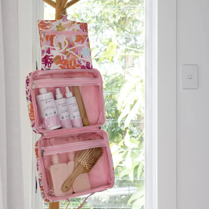 A pink and white floral hanging toiletry organizer bag with two open compartments, shown hanging on a wooden door hook with skincare bottles and a hairbrush inside, against a bright window background.