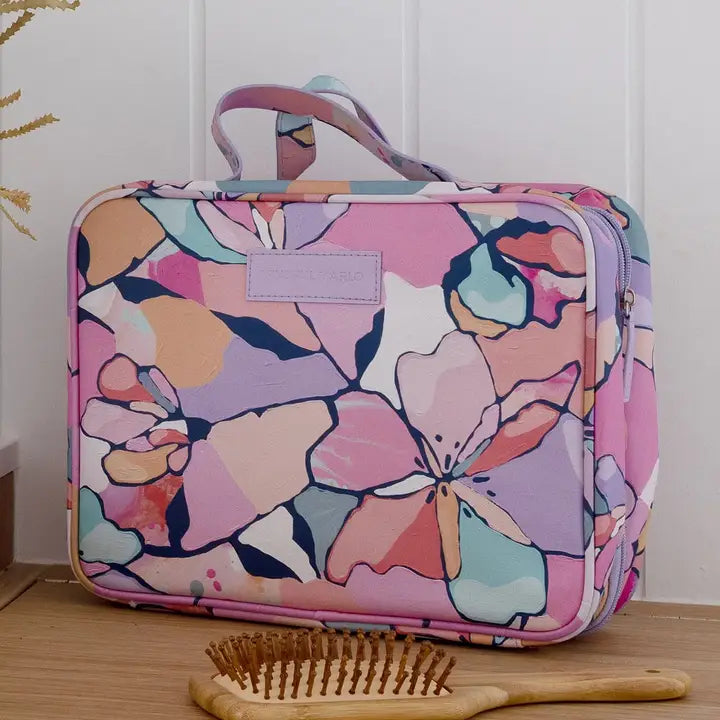 A rectangular toiletry bag with a colorful floral pattern in pink, purple, blue, and peach, shown on a wooden surface with a wooden hairbrush in front. 