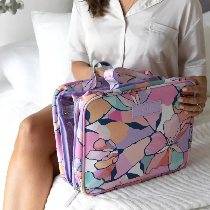 A person in a white pajama shirt holds a colorful floral toiletry bag while sitting on a white bed. The bag has a pink, purple, and peach floral pattern with a lavender brand patch on the front.