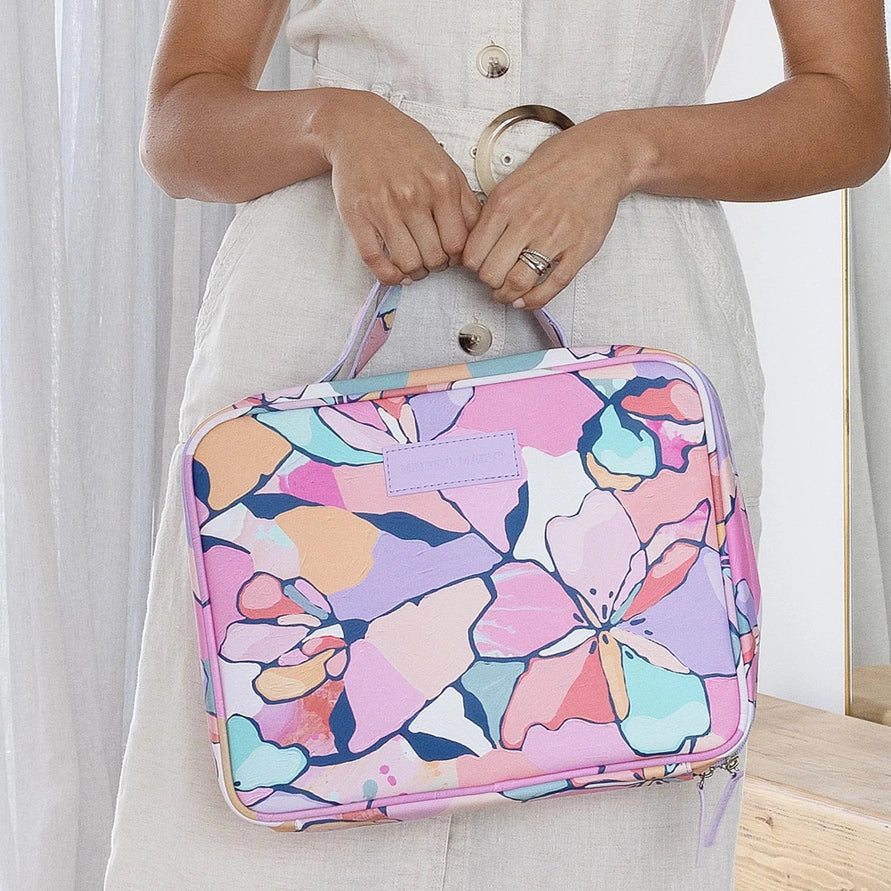A woman in a light-colored dress holds a rectangular handbag with a colorful floral pattern in pink, purple, blue, and orange. The bag has a top handle and a zip-around closure.