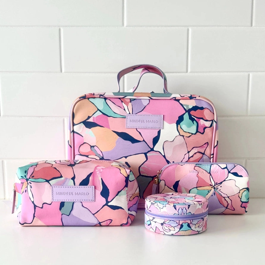 A matching toiletry bag set by Monstera Milano with a colorful floral pattern in pink, purple, orange, and teal, displayed on a white surface against a white subway tile wall.