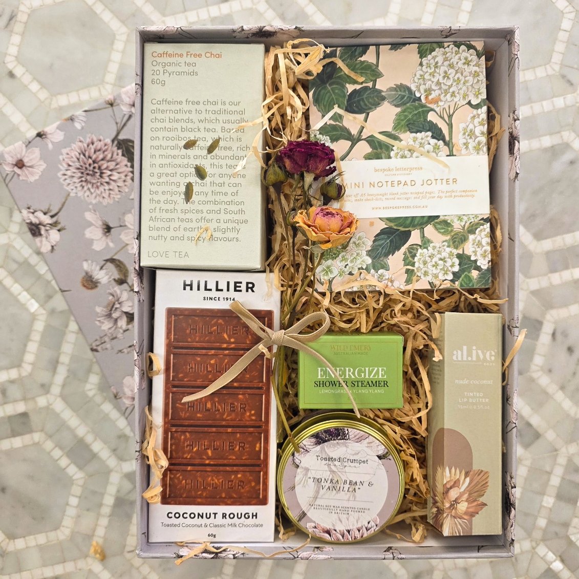 Gift set with tea, chocolate, and other items in a decorative box on a patterned surface.