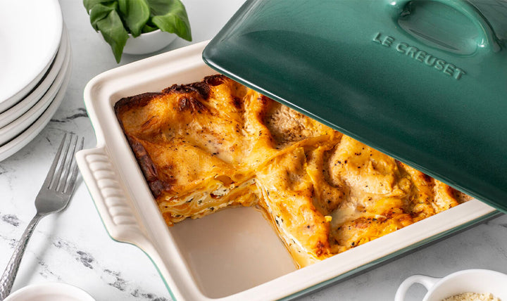 Lasagna in a Le Creuset dish on a table with plates and a plant in the background