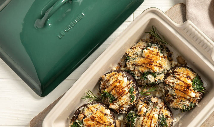 Baked mushrooms with herbs in a ceramic dish next to a green Le Creuset dish.