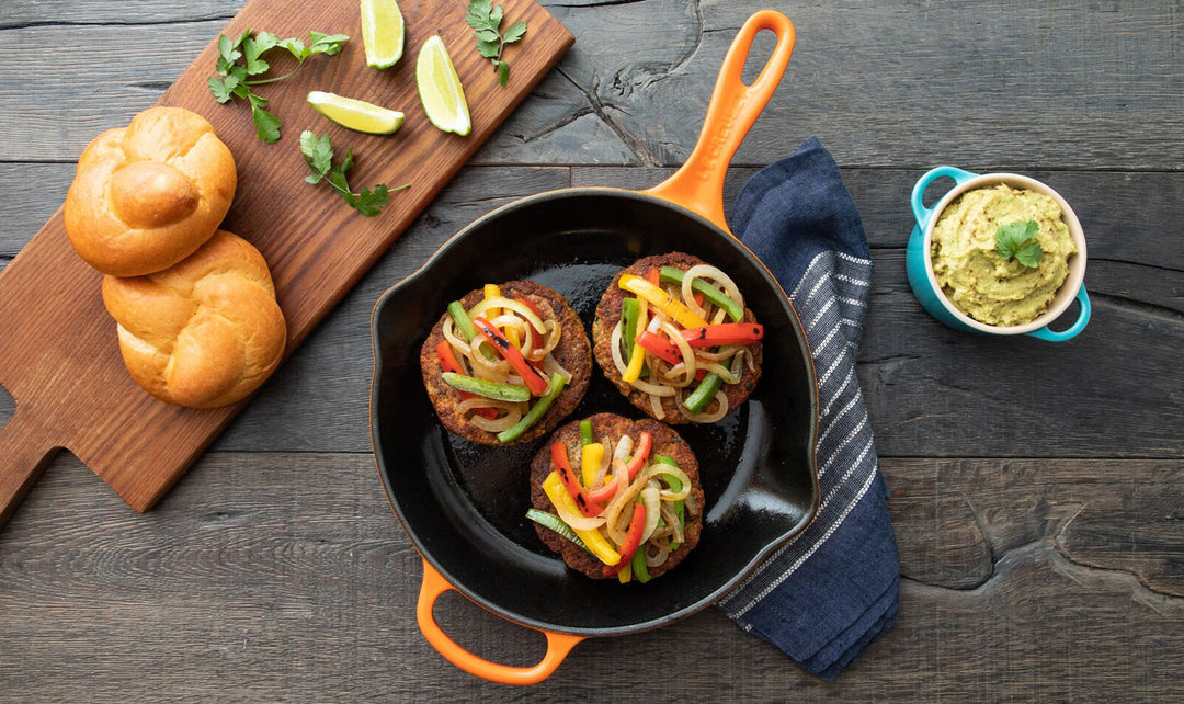 Skillet with stuffed burgers topped with peppers and onions, surrounded by bread and guacamole on a wooden surface.