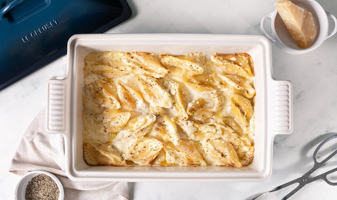 Baked pasta dish in a white casserole dish on a light gray surface with a Le Creuset pot in the background.