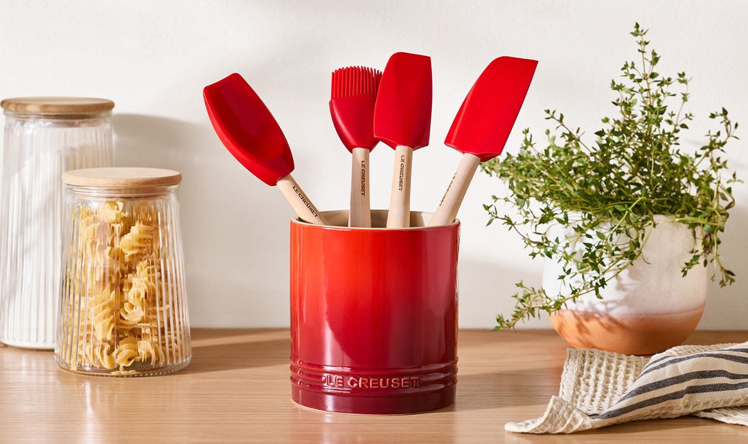 Red utensil holder with spatulas on a kitchen counter next to jars and a plant.