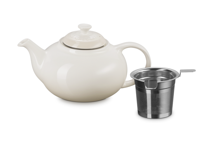 Le Creuset cream ceramic teapot with a metal infuser basket on a white background