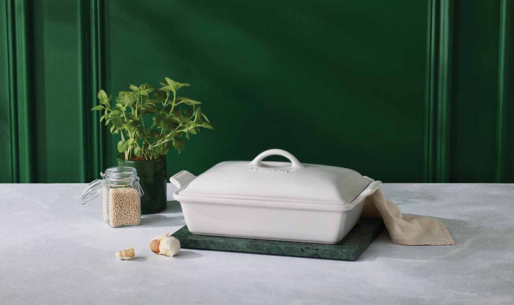 White ceramic dish with lid on a green surface with a green plant and jar in the background.