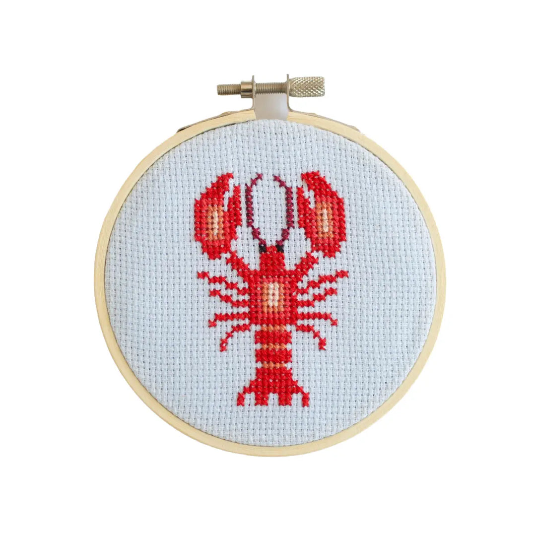Cross-stitched lobster design on fabric in a wooden hoop against a white background