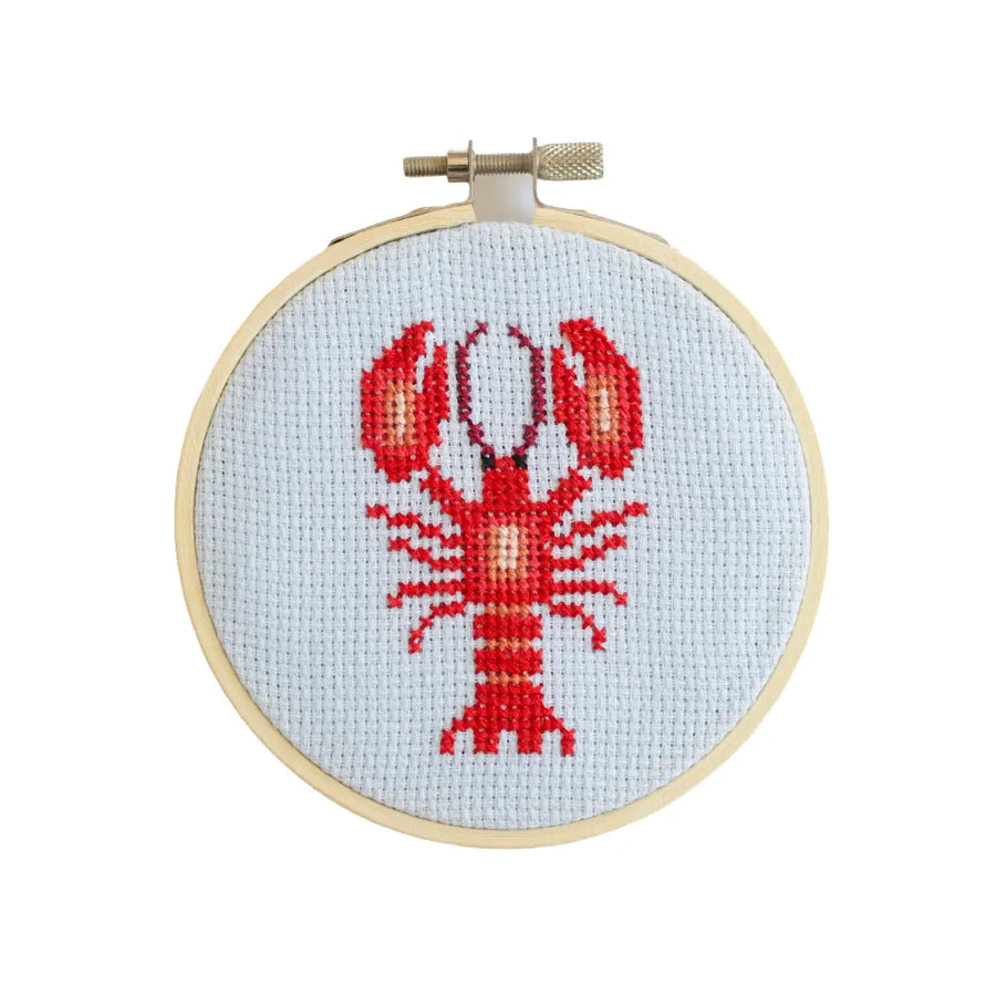 Cross-stitched lobster design on fabric in a wooden hoop against a white background