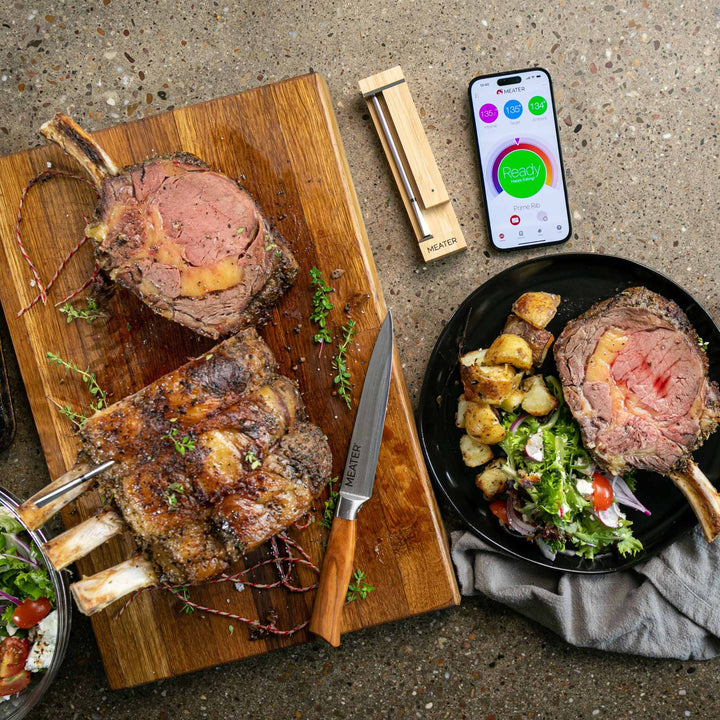 Two roasted lamb chops on a wooden cutting board with a smartphone displaying a cooking app, on a concrete surface.