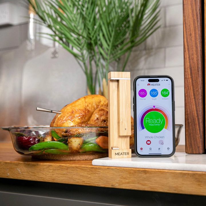 Smart meat thermometer with smartphone app displayed on a kitchen counter next to a roasted chicken.