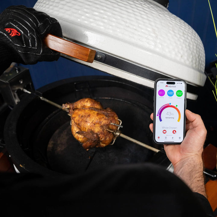 Person grilling a chicken on a barbecue with a smartphone app displayed on a screen.
