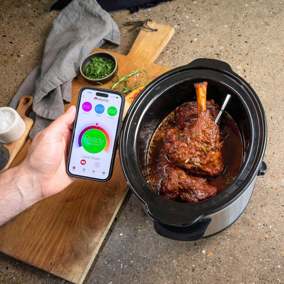 Person holding a smartphone with a cooking app next to a slow cooker with food inside.