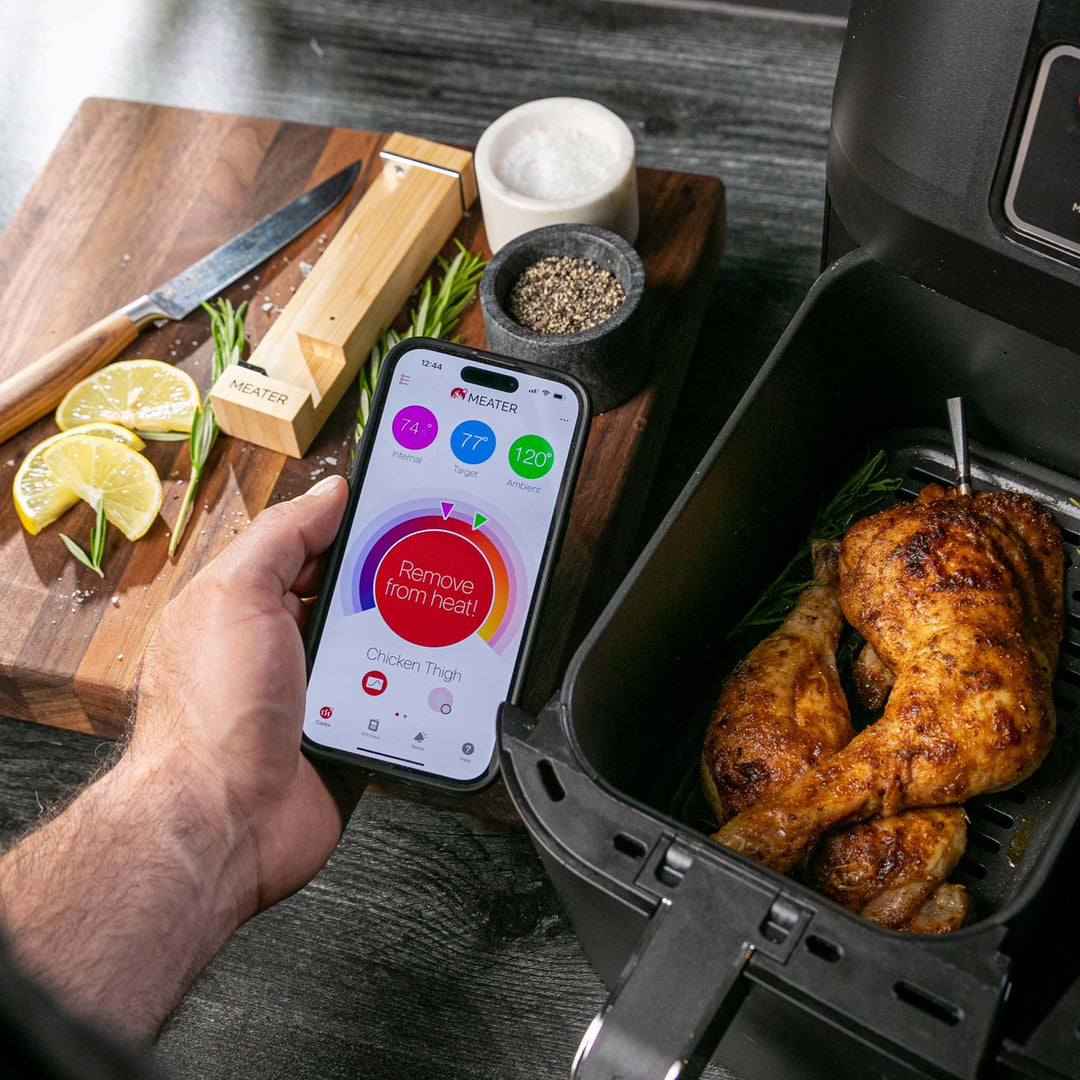 Person using a smartphone app to control an air fryer with roasted chicken inside, on a kitchen counter.
