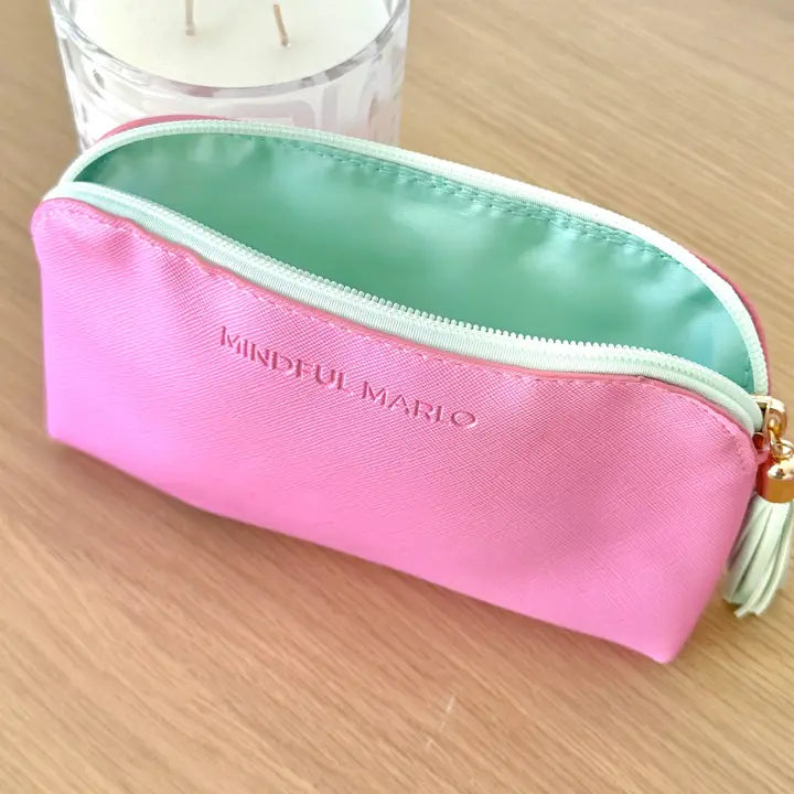 A pink zippered pouch with a mint green interior and a gold tassel zipper pull, placed on a light wood surface. The brand name &