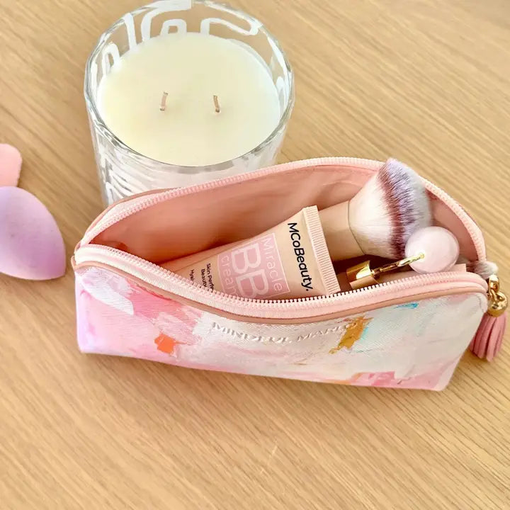 A pink cosmetic pouch with a pastel watercolor print sits open on a light wood surface, containing makeup items like a tube and brushes. A lit candle and soft pink props are visible in the background.