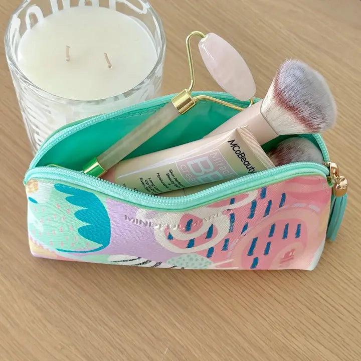 A pastel patterned cosmetic pouch with a turquoise zipper, shown open on a light wood surface with makeup items inside, including a brush, tube, and roller tool. A candle is partially visible in the background.