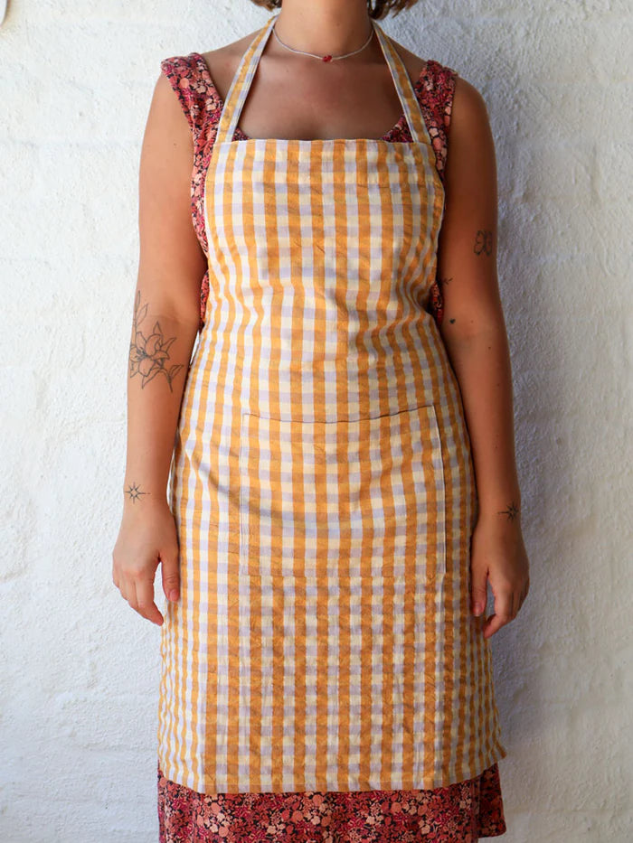 Person wearing a yellow checkered apron over a floral dress against a white wall.