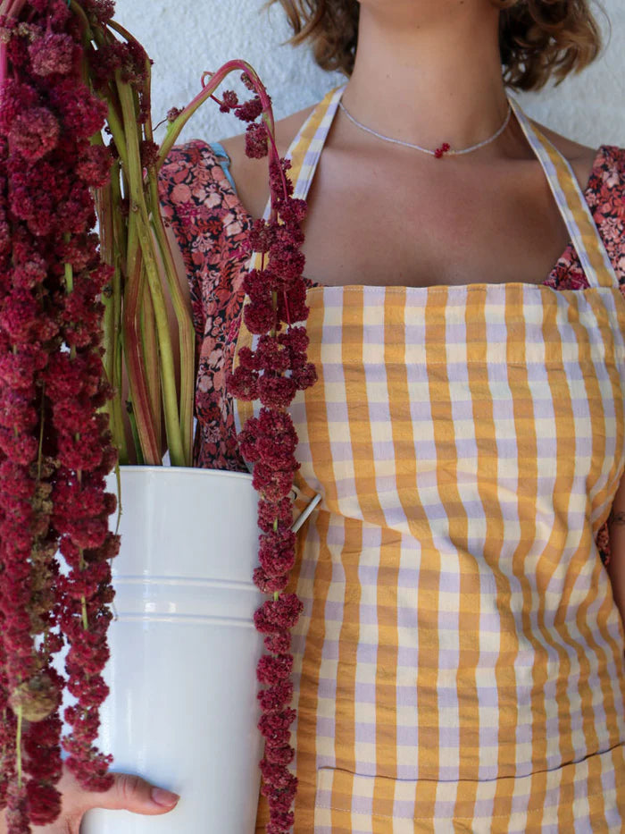 Person wearing a yellow and white checkered apron holding a bucket with red plants.