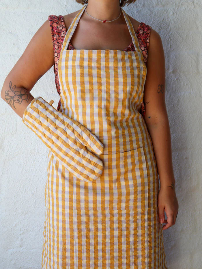 Person wearing a yellow and white checkered apron over a floral dress against a white wall.