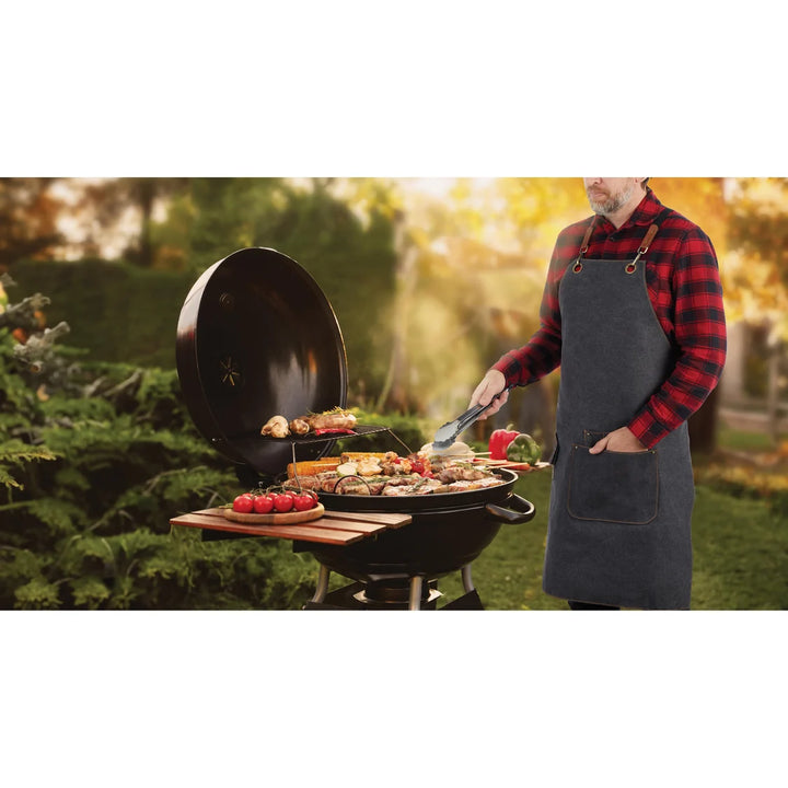A man wearing a dark gray kitchen apron over a red and black plaid flannel shirt grills food outdoors on a black kettle-style barbecue grill in a garden setting.