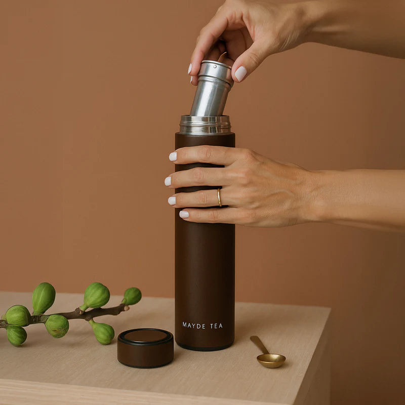 Hand holding a brown thermos with 'Mayde Tea' branding on a wooden surface with green leaves and a spoon.