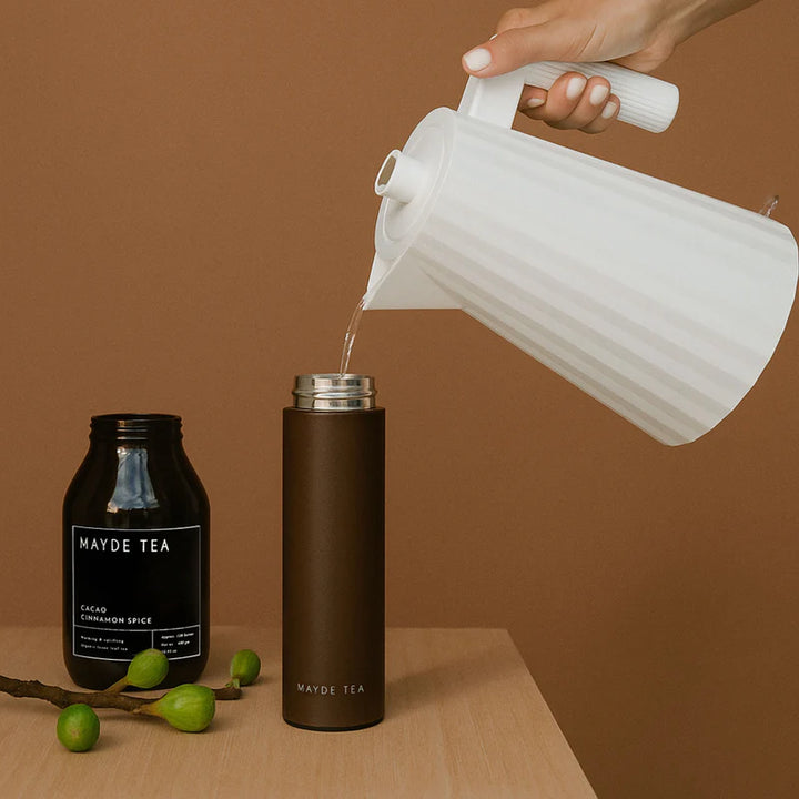 Person pouring liquid into a brown thermos with Mayde Tea packaging on a wooden surface and brown background.