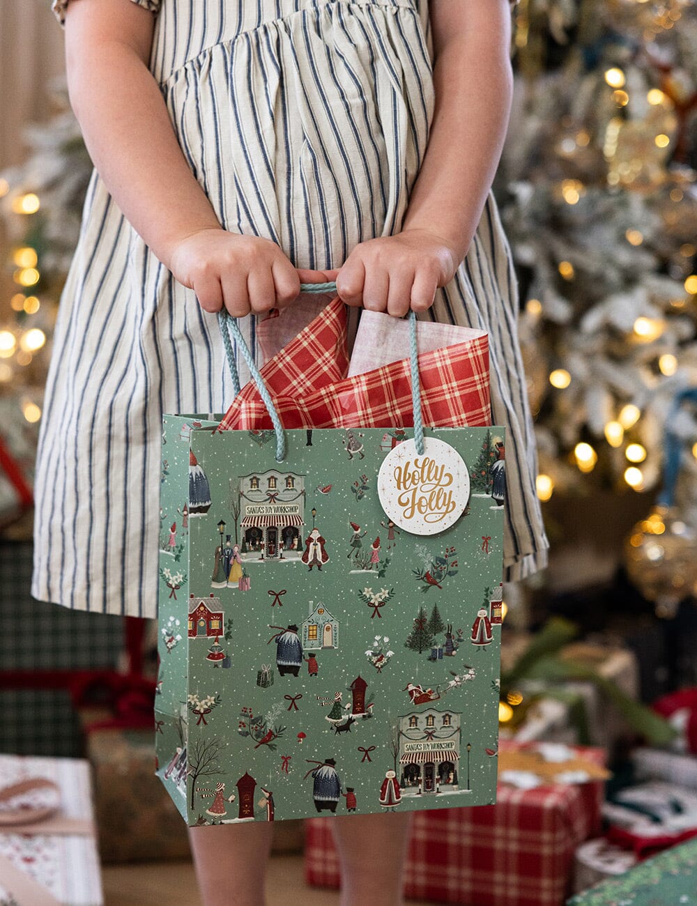 Child holding a festive gift bag with a Christmas tree and presents in the background