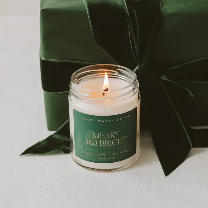 Candle in a jar with a green label reading 'Merry and Bright' surrounded by green leaves on a light background.