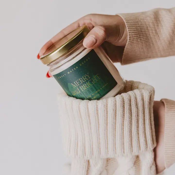 Hand holding a candle labeled 'Merry & Bright' with a person wearing a beige sweater.