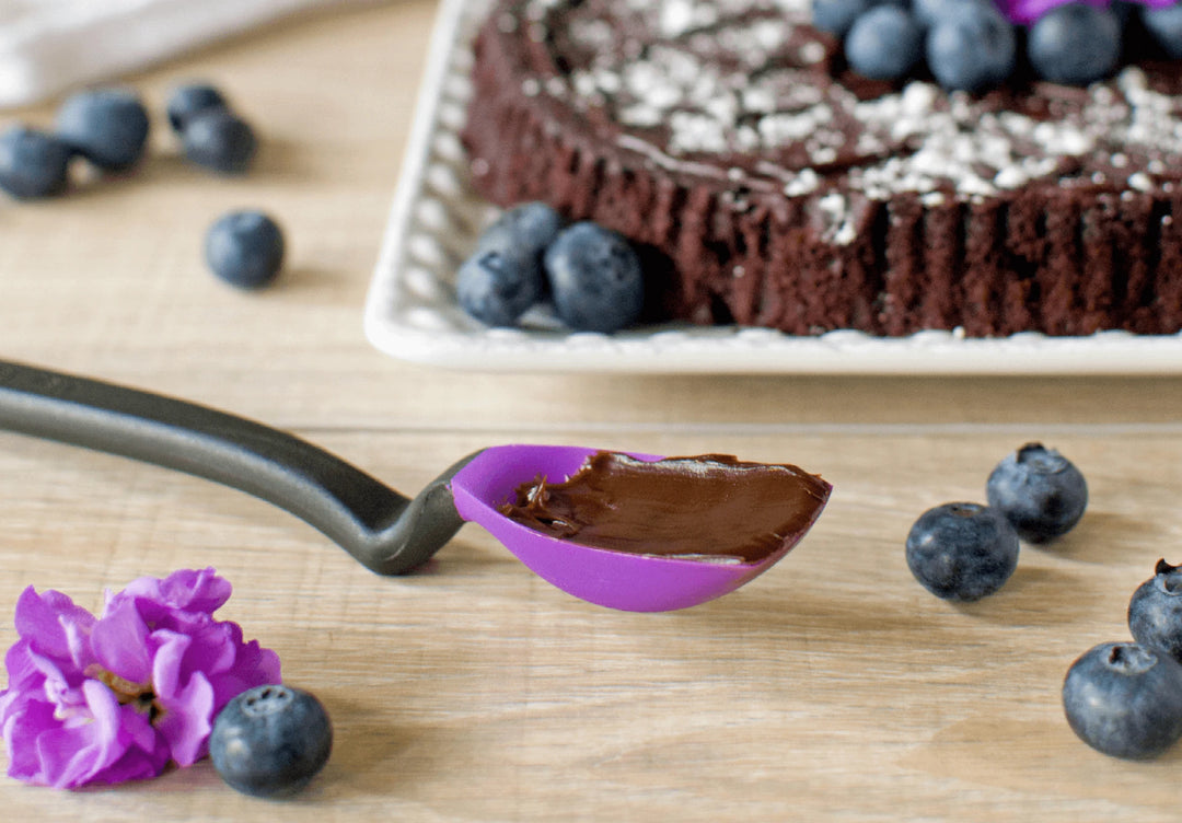 Chocolate tart with blueberries on a wooden surface, surrounded by a spoonful of chocolate and purple flowers.