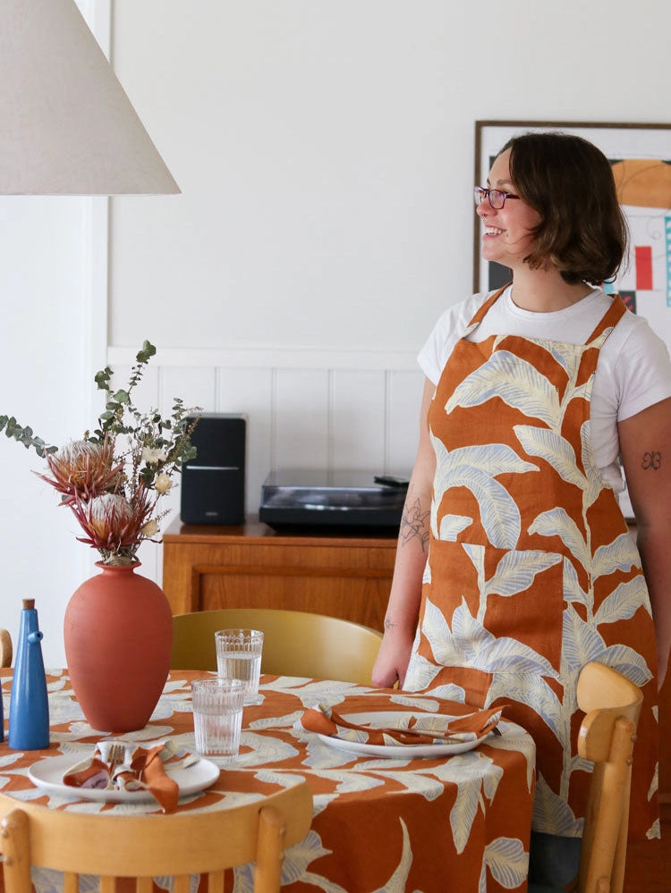 Person wearing a patterned apron standing by a dining table with a vase and decor items.