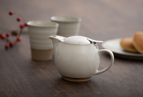 Ceramic teapot with a spoon on a wooden table with two cups and a plate of bread in the background.